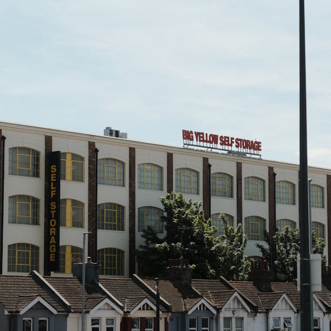 View of a large, multi-storey building with a white exterior and multiple yellow-framed windows, housing a self-storage facility named 'Big Yellow Self Storage' in red signage at the top. A vertical black sign with yellow lettering reading 'Self Storage' is mounted on the building's side. In the foreground, there are smaller residential-style houses with brown tiled roofs, white gable ends, and chimneys, positioned along a street. A row of trees partially obscures the lower part of the building, and a streetlamp is visible on the right side of the image. The scene is captured in daylight, with a partly cloudy sky overhead, reflecting a typical urban environment suitable for home relocation and furniture transport services provided by companies like Man with Van Brentford, specialising in removals and moving logistics.