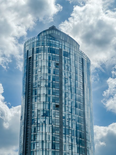 Photograph of a modern high-rise office building with a curved glass facade, reflecting the blue sky and white clouds above. The building's exterior features numerous windows framed by metallic structures, creating a sleek and contemporary appearance. The top of the building is rounded, and the reflection of the sky gives it a bright, airy feel. This urban architecture image aligns with themes of property and building management, potentially relevant to house removals and relocation logistics. The picture is taken from a low angle, emphasizing the tower's height against the backdrop of partially cloudy skies. In the context of home relocation, the image subtly highlights the importance of planning for moving into or out of properties within such urban environments, where careful logistics and transportation of furniture and belongings are essential. The visual connection to property and building structures reinforces the services offered by [COMPANY_NAME], located at [PAGE_URL], focusing on removals and moving services in Brentford.