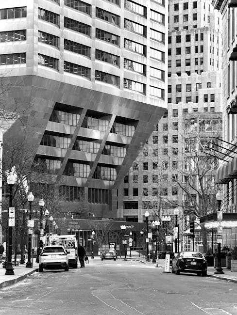 Black and white photograph of a busy city street in an urban area, featuring a large modern building with a distinctive overhanging section supported by angled supports. The building has numerous rectangular windows and appears to be constructed with concrete or stone. Several parked cars are visible along the street, with a few pedestrians walking on the pavement. In the foreground, there are streetlamps, traffic signs, and trees without leaves, indicating a colder season. The scene captures the typical environment surrounding a commercial or office district with high-rise buildings. Man with Van Brentford often operates in areas with such urban infrastructure, supporting home and office removals, packing, and furniture transport, ensuring efficient logistics during house relocations or business move projects.