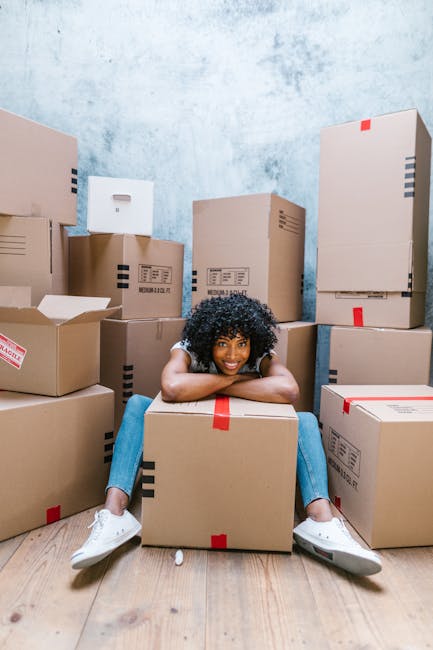 A young woman with curly black hair, dressed in a white t-shirt, blue jeans, and white sneakers, is seated on a light wooden floor inside a room filled with stacked cardboard moving boxes of various sizes, some sealed with red and black packing tape. She is leaning forward with her arms resting on a large box in front of her and smiling while looking directly at the camera. The room has a plain, textured light blue wall in the background. The boxes surround her, some partially open revealing packing materials, indicating a home relocation process supported by Man with Van Brentford. The scene captures a moment during packing or loading for furniture transport, with the focus on the organised array of boxes and the user's relaxed pose, symbolising efficient packing and moving logistics.