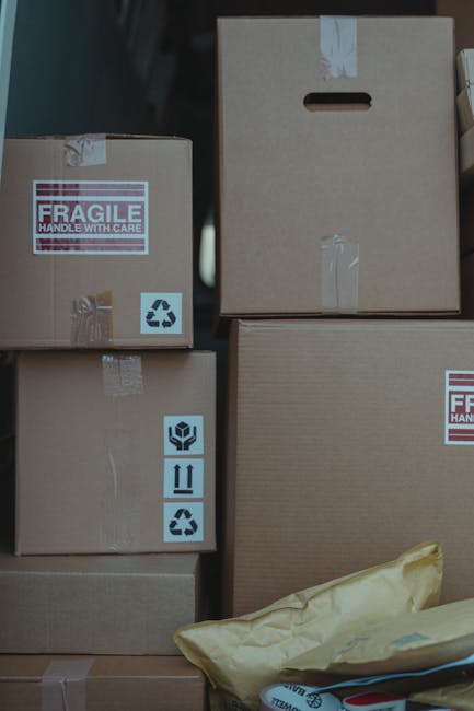 A close-up view of several stacked cardboard moving boxes inside a property, with some boxes sealed with clear packing tape and marked with recycling and handling symbols. One box features a prominent red and white 'Fragile Handle with Care' label. The boxes are of various sizes, arranged on a surface or shelving unit, with a yellow parcel or packing bag in front. The background suggests an indoor environment, possibly a storage area or room prepared for home relocation, and parts of a door or doorway are visible behind the boxes. This scene illustrates typical packing materials and the careful arrangement involved in furniture transport and moving logistics, as part of an emergency same-day removal service by Man with Van Brentford.