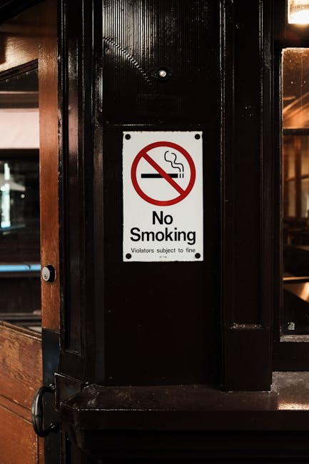 A close-up image of a black wooden partition inside a property, featuring a white rectangular sign with a red circle and slash, indicating 'No Smoking' with a cigarette icon inside. The sign includes the text 'No Smoking' and a small note beneath stating 'Violators subject to fine.' The setting appears to be part of a home or public establishment with warm ambient lighting visible in the background through glass panels, possibly a bar or lounge area. This image reflects the importance of adhering to no-smoking policies during home relocation or furniture transport activities to ensure safety and compliance in the moving process. The black wood surfaces and the sign are clearly visible, highlighting areas where removals teams, such as those from Man with Van Brentford, operate when preparing properties for moves or transporting items safely.