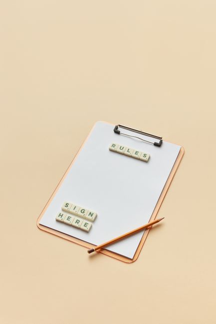 A white clipboard with a metal clip at the top rests on a light beige surface. On the clipboard, there are two sets of lettered Scrabble tiles; the first set spells 'RULES' positioned horizontally at the top, and the second set spells 'SIGH HERE' placed towards the bottom. A thin, orange-brown pencil lies diagonally across the lower right corner of the clipboard, with its tip pointing toward the tiles. The background is neutral, and the overall scene appears to be arranged for note-taking or planning, subtly relating to household organisation and moving preparations as seen in house removals and furniture transport services provided by Man with Van Brentford.