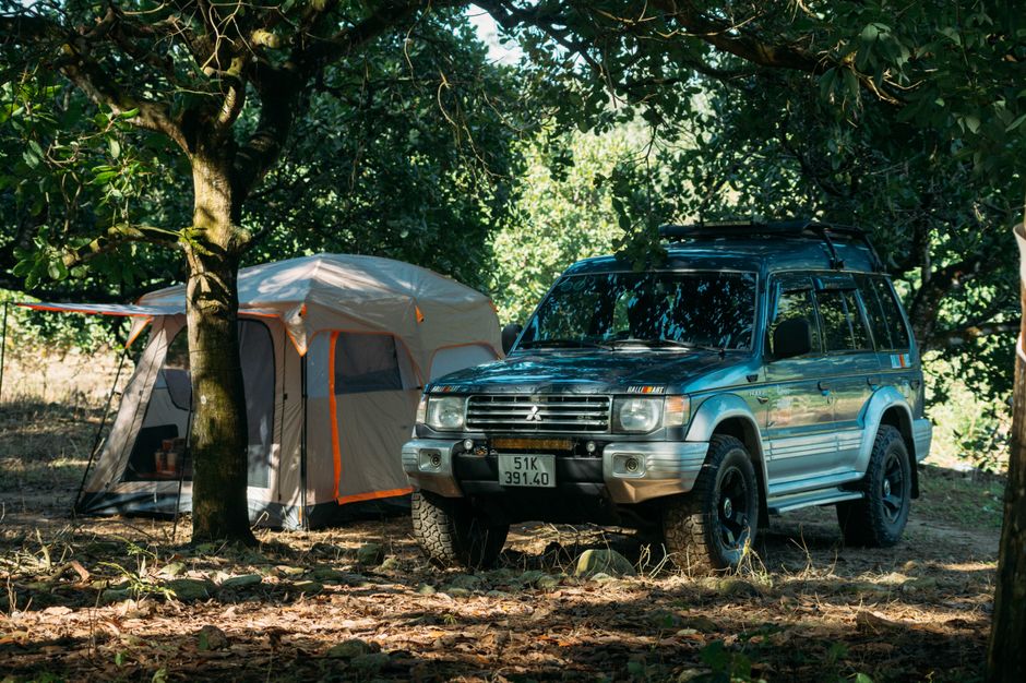 A blue Mitsubishi Pajero off-road vehicle parked on uneven ground within a wooded area, surrounded by tall trees with green foliage providing partial shade. The vehicle has a roof rack and appears to be part of a home relocation or outdoor adventure, with visible dirt and mud on its tires and lower body. Nearby, there is a beige camping tent with orange accents made of fabric and mesh panels, positioned next to a tree, suggesting outdoor living accommodation typical of lifestyle or camping activities. The tent is partially unzipped, revealing interior gear, possibly related to packing or outdoor gear transport. The scene is illuminated by natural daylight, with dappled sunlight filtering through the leaves, highlighting the outdoor environment. This setting illustrates the process of loading or unloading outdoor equipment during a move or outdoor trip, aligning with the theme of transporting belongings and outdoor packing as part of a professional removals or relocation service, such as offered by Man with Van Brentford.