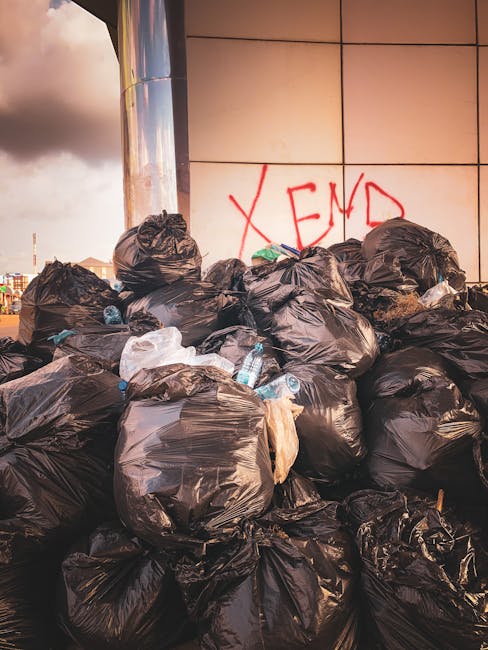 A large pile of black trash bags filled with bulky household waste, including plastic bottles and packaging, is stacked outdoors next to a tiled wall with red graffiti reading 'XEND.' The bags are placed on a pavement surface near the entrance of a property, with some bags slightly torn or deformed, indicating recent collection or disposal. Behind the bags, part of a metallic structure or column is visible, and the sky shows a mix of clouds and sunlight, suggesting late afternoon or early evening. The scene exemplifies waste disposal during a home removal or clearance process, with the bags ready for collection by waste services or for transport during a furniture or property relocation, as handled by companies like Man with Van Brentford.
