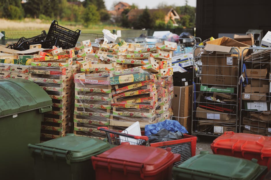 Outdoor scene showing a large collection of household waste and recyclable materials prepared for disposal or removal, including multiple stacks of cardboard boxes filled with empty produce trays labeled 'fresh fruits,' arranged on a pavement area. Several black plastic crates and metal storage racks are visible, holding various items such as flattened cardboard, packaging boxes, and miscellaneous debris, some wrapped in brown paper or plastic wraps. In the foreground, there are multiple large plastic wheelie bins in green and red, positioned along the sidewalk or driveway, ready for collection or transportation. Part of a red shopping trolley and a blue plastic bag are also seen near the bins, indicating loading activity. The background features a residential neighborhood with houses and greenery, suggesting the scene occurs during a house relocation or decluttering process. The environment is well-lit with natural daylight, and the setting reflects typical packing or disposal preparations carried out by professional removals services like Man with Van Brentford, supporting efficient furniture transport and packing logistics related to home relocations and waste disposal in Brentford.