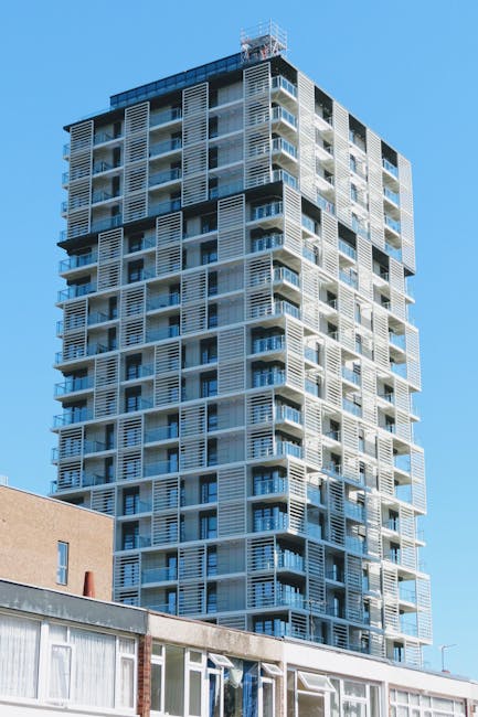 A high-rise residential building under construction, featuring multiple floors with large glass windows and white external metal louvered shading panels. The building is situated in an urban environment with a clear blue sky overhead. In the foreground, part of an older, lower-rise building with a flat roof and visible window frames is shown, providing context for the nearby infrastructure. The construction site is well-lit with natural daylight, highlighting the architectural details and modern design. This image exemplifies ongoing development in the area, often associated with urban home relocation and property improvements. Man with Van Brentford offers professional removals and furniture transport services for residents and companies relocating within or around Brentford, supporting smooth moving processes within such high-rise developments or nearby properties.