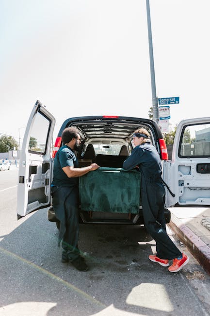 Two movers from Man with Van Brentford are engaged in a home relocation task, loading a large, dark green upholstered piece of furniture into the open rear of a white delivery van parked on a street. One mover, wearing a blue uniform and glasses, is carefully lifting the item while the other, dressed similarly, provides assistance from the opposite side. The van is situated on a paved area beside a curb with a parking sign and a lamppost nearby, under clear daylight conditions. Inside the van, the cargo space appears spacious, ready to accommodate furniture or boxes, typical of packing and moving operations. The background includes a street scene with trees and buildings, indicating an urban environment in Brentford. This process reflects professional furniture transport and logistics involvement by [COMPANY_NAME], highlighting efficient loading procedures essential in residential moves.