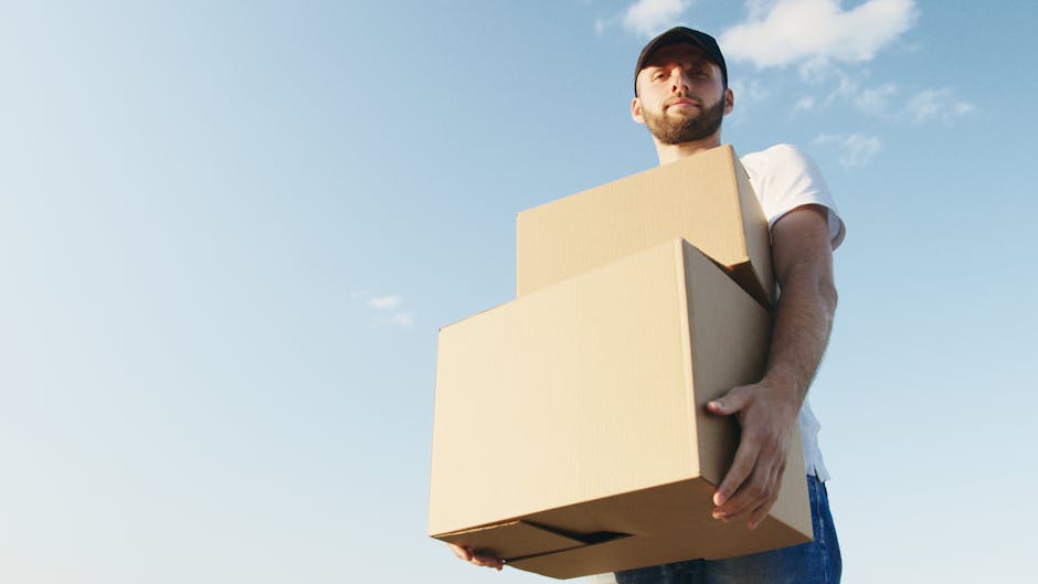 A man with a beard and wearing a black cap, white t-shirt, and blue jeans is standing outdoors against a blue sky with a few scattered clouds, holding three cardboard boxes of varying sizes stacked one on top of the other. He appears to be preparing for a home relocation, as part of a furniture transport or moving process. The boxes are sealed with clear packing tape, and he is supporting them securely with both hands. In the background, there is a paved area or driveway, suggesting a loading zone adjacent to the property. The scene is well-lit with natural daylight, emphasizing the practical nature of packing and moving activities. As part of the services offered by Man with Van Brentford, this image captures the initial stages of loading household items for a local removal or furniture transport, illustrating careful handling of packed belongings ready for delivery to a new home.
