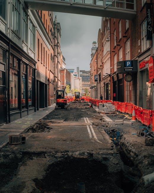 The photograph depicts a narrow urban street undergoing roadworks, with uneven asphalt and excavation areas, surrounded by multi-storey brick and stone buildings on both sides. In the foreground, orange plastic safety barriers line the edge of the construction zone, and construction equipment such as small excavators and marked vehicles are visible further down the street. The street is lined with signage, including parking signs, and displays a partially covered pedestrian walkway overhead. Natural daylight illuminates the scene, providing clear visibility of the ongoing work. This setting reflects a typical city centre undergoing infrastructure improvements, which can impact home relocation or furniture transport logistics. Man with Van Brentford offers removals and moving services in such environments, managing the loading process carefully to navigate narrow access routes and construction zones.