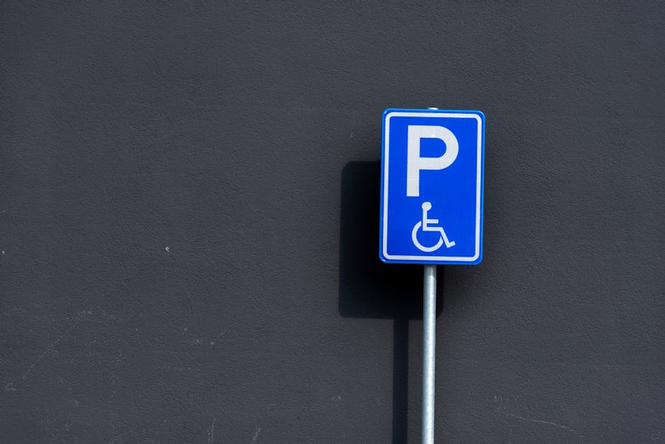 A close-up view of a blue parking sign with a white 'P' and a wheelchair symbol, mounted on a metallic pole. The sign is positioned against a smooth, dark grey wall in an outdoor setting. In the background, faint shadows of the sign are cast onto the wall. The image captures the sign at a slight angle, indicating its placement on a pavement or parking area adjacent to a building. The lighting is natural, with even illumination highlighting the sign's clear markings. This scene relates to vehicle parking and accessibility, relevant to house removals and moving logistics services provided by Man with Van Brentford, especially when considering parking permits or restrictions during home relocation or furniture transport.