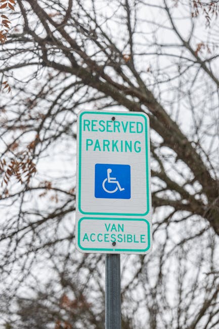 A rectangular reserved parking sign with a green border, mounted on a metal pole against a background of leafless tree branches and an overcast sky. The sign displays the wheelchair symbol in blue and white, indicating disabled parking privileges, with the words 'RESERVED PARKING' at the top and 'VAN ACCESSIBLE' at the bottom in green text. This sign is positioned on the pavement near a property where a house moving process is underway. Although the image does not show moving equipment or vehicles directly, it is relevant within the context of logistics for home relocation services by Man with Van Brentford, which often involves navigating parking restrictions and obtaining permits for efficient furniture transport and packing and moving activities.