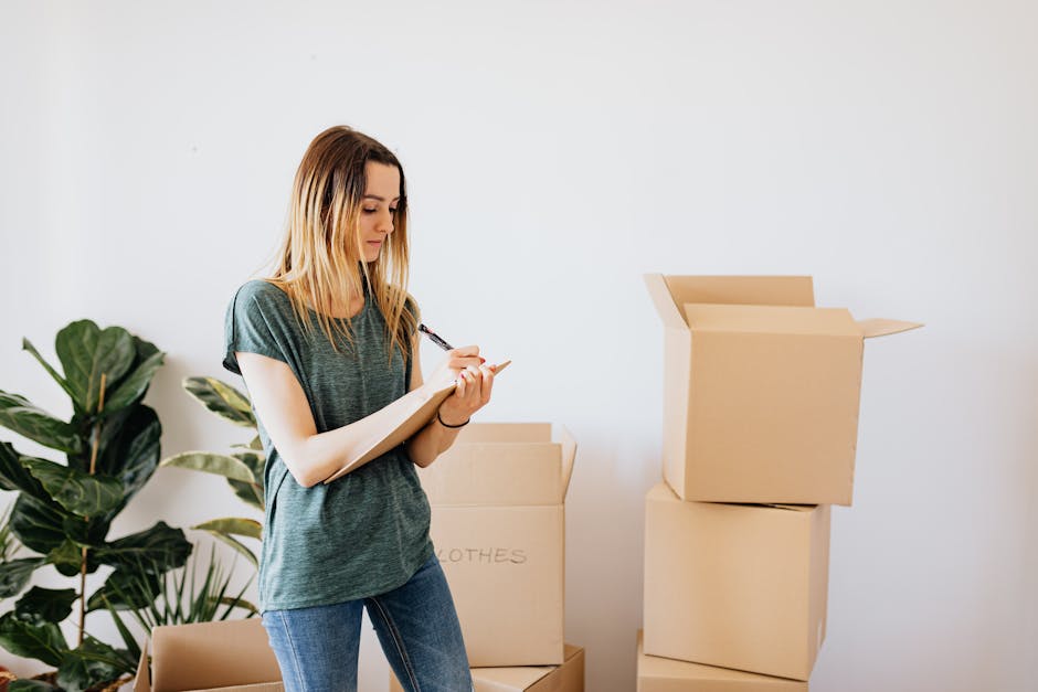 A young woman with long blonde hair, dressed in a casual grey t-shirt and blue jeans, stands inside a property during a house move, taking notes on a clipboard with a pen. She is surrounded by several
